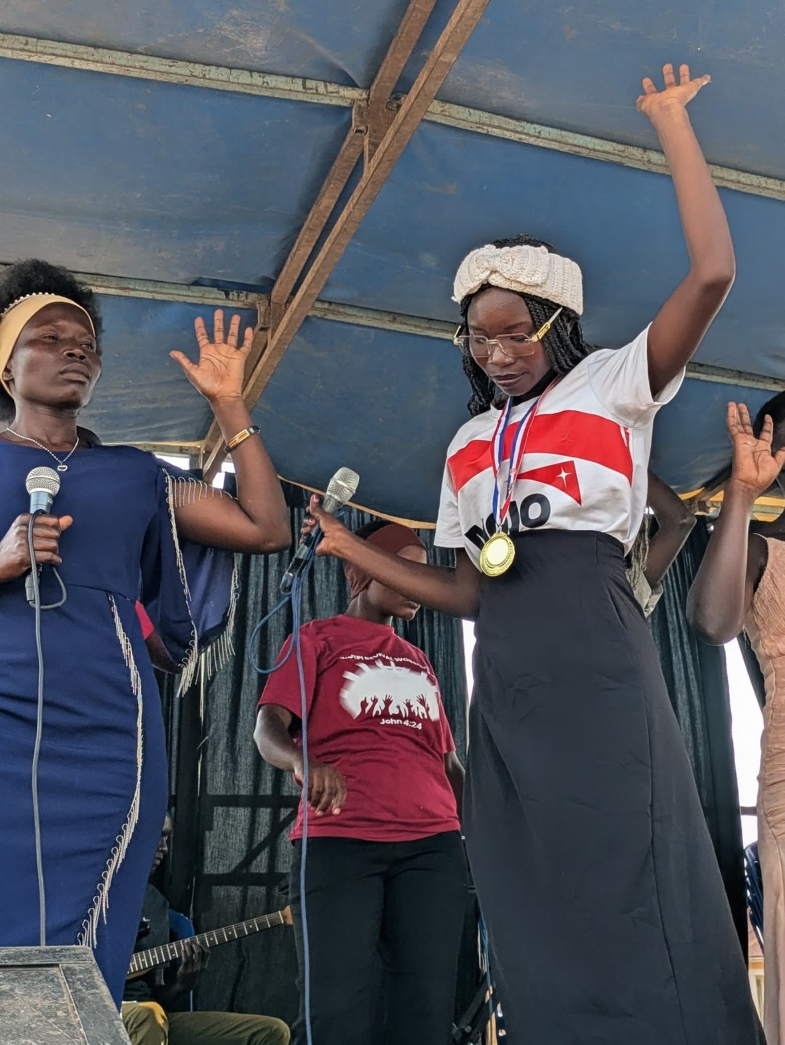 Two young women speaking on stage at a community gathering in Rhino Camp