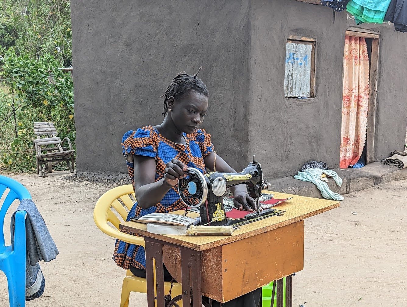 A woman sewing clothes at a treadle machine outside a house in Rhino Camp